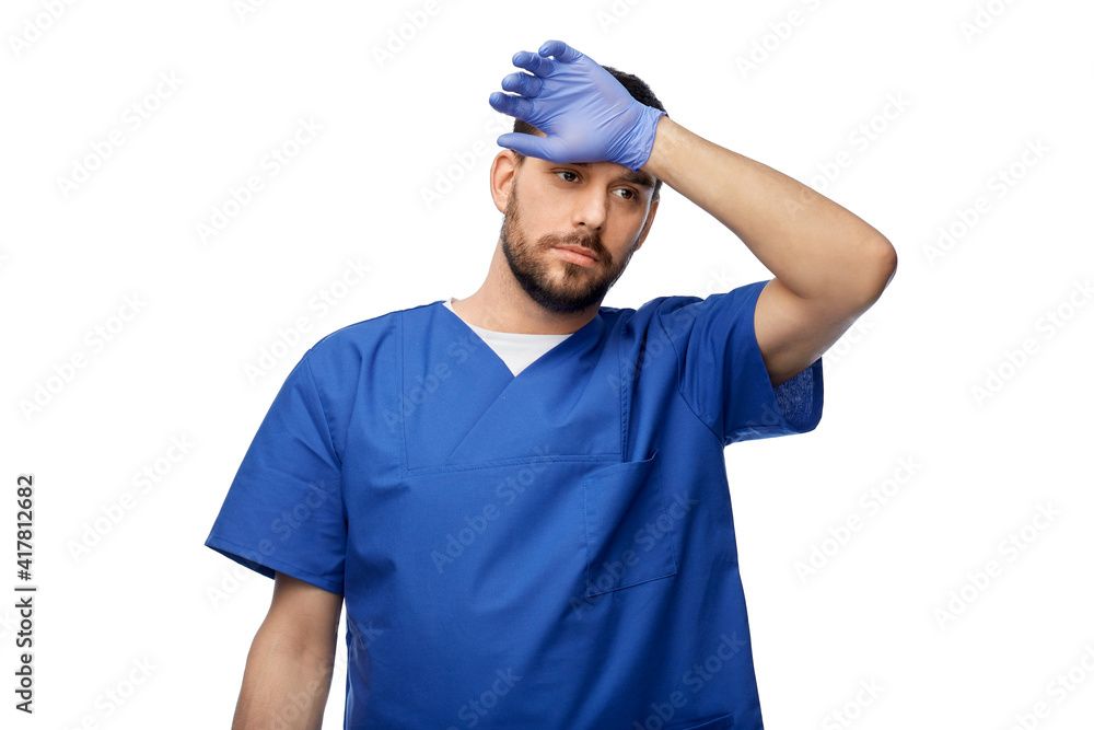 healthcare, profession and medicine concept - tired doctor or male nurse in blue uniform and medical gloves on over white background