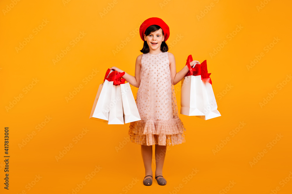 Fototapeta premium Little french girl holding paper shopping bags. Studio shot of female kid in dress and red beret.