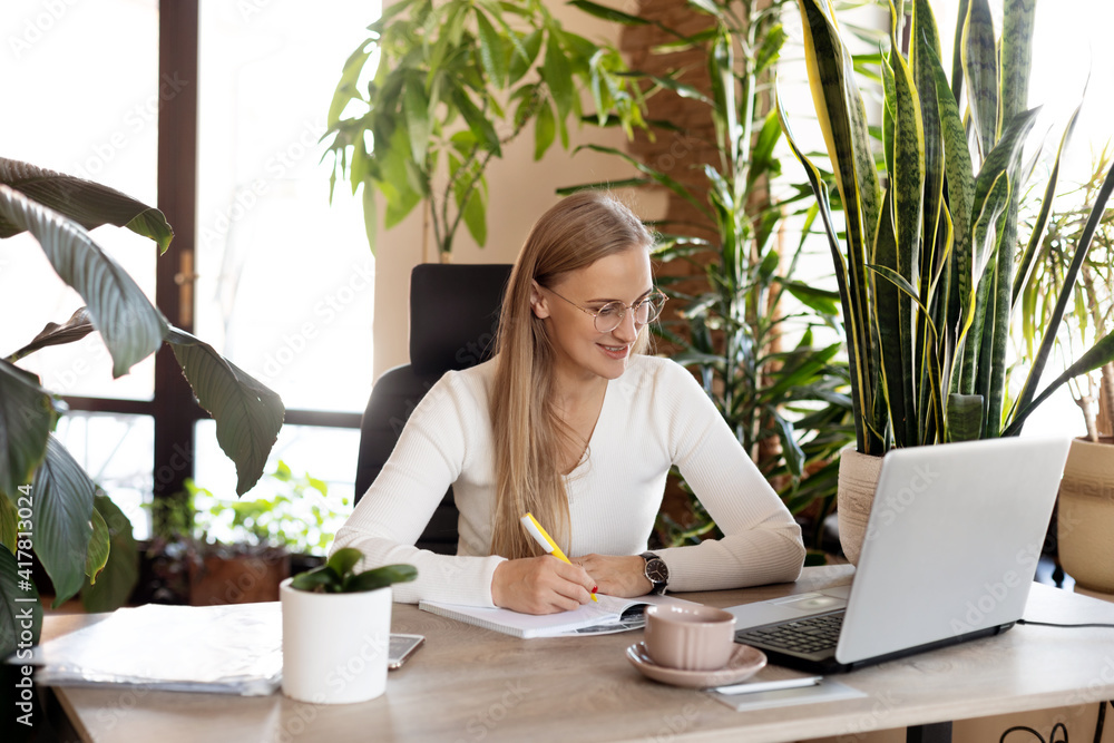 A beautiful business woman works at a laptop in the office and makes notes in a notebook. Business, online business, work in the office A beautiful business woman works at a laptop in the office and makes notes in a notebook. Business, online business, work in the office