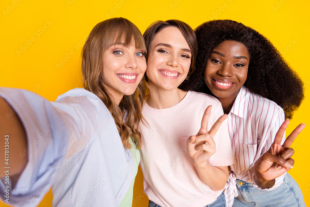 Portrait of three charming cheerful ladies make selfie fingers show v-sign isolated on yellow color background Portrait of three charming cheerful ladies make selfie fingers show v-sign isolated on yellow color background