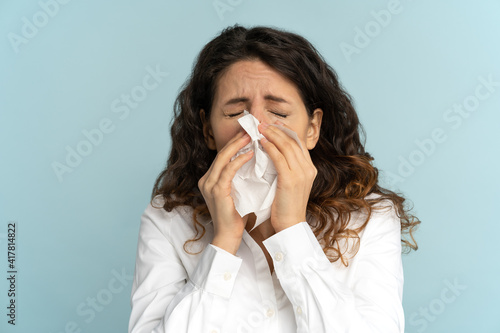 Close up studio portrait of young office employee woman in white blouse with tissue, sneezing, suffering from seasonal allergy, isolated on blue background. Rhinitis, symptoms of flu virus concept.