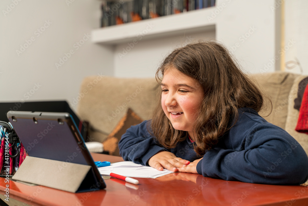 a little girl smiles happily as she communicates via video conference with her teacher using a tablet while sitting on the sofa at home to do her homework. Online education concept a little girl smiles happily as she communicates via video conference with her teacher using a tablet while sitting on the sofa at home to do her homework. Online education concept