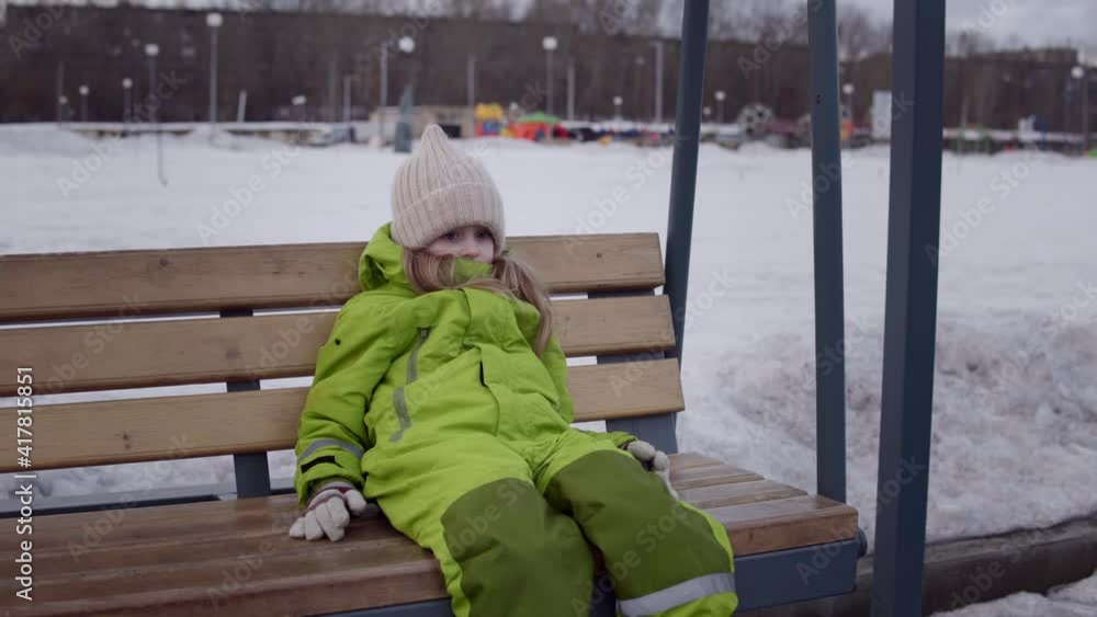 A freezing child in a beige knit hat and green snowsuit swings on a swing with a wooden seat in a snow-covered park against the backdrop of a children's playground A freezing child in a beige knit hat and green snowsuit swings on a swing with a wooden seat in a snow-covered park against the backdrop of a children's playground