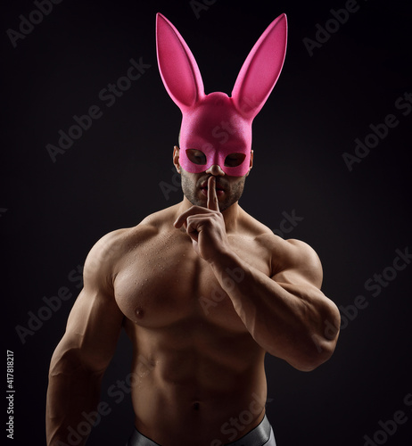 Obraz na płótnie Portrait of shirtless handsome muscular man, athlete with perfect built body wearing pink rabbit, bunny mask stands with naked chest and gesturing silence, secret sign with finger on dark background w salonie