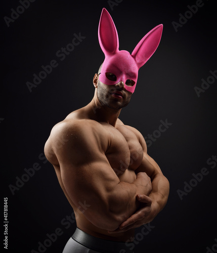 Obraz na płótnie Portrait of shirtless handsome muscular man, athlete with perfect built body wearing pink rabbit mask standing naked sideways to camera demonstrating his biceps and chest over dark background w salonie