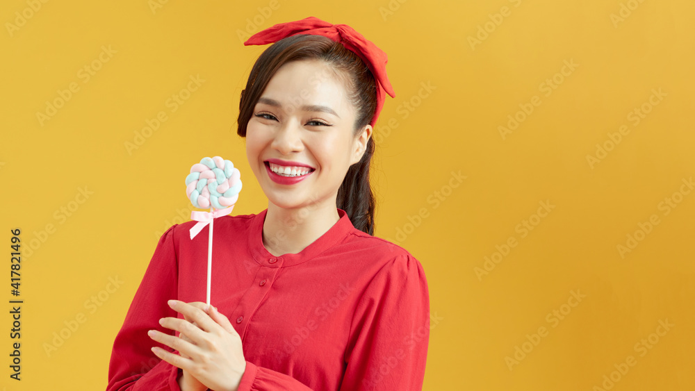 Pleasant looking cheerful woman poses with delicious lollipop enjoys leisure time against yellow background Pleasant looking cheerful woman poses with delicious lollipop enjoys leisure time against yellow background