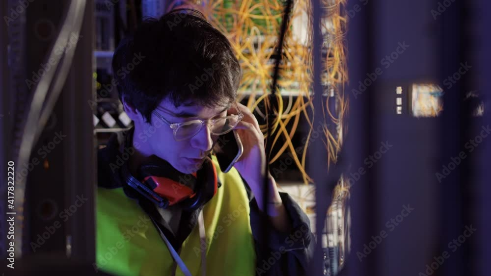 Medium shot of young Asian IT engineer wearing special uniform and glasses standing in data center, making photo of wires using smartphone camera and then sending photo to manager Medium shot of young Asian IT engineer wearing special uniform and glasses standing in data center, making photo of wires using smartphone camera and then sending photo to manager