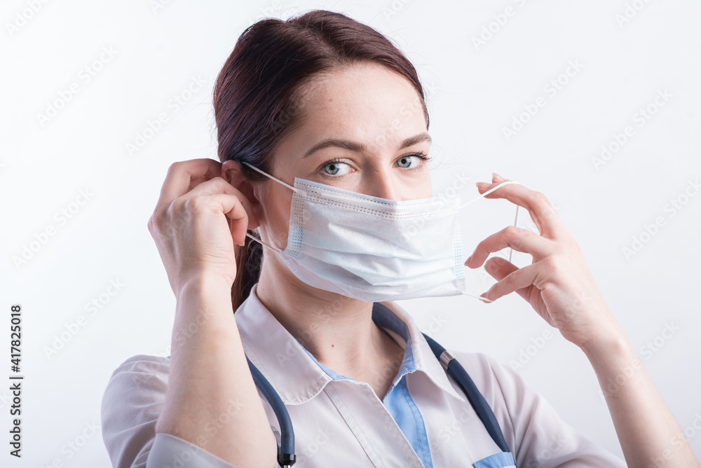Portrait of a female doctor in a white uniform who puts on a medical mask in the studio on a white background Portrait of a female doctor in a white uniform who puts on a medical mask in the studio on a white background
