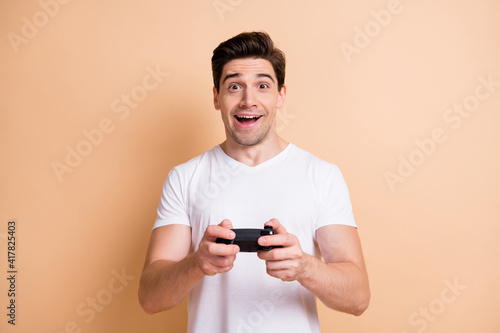 Portrait of impressed brunet man playstation wear white t-shirt isolated on beige color background © deagreez