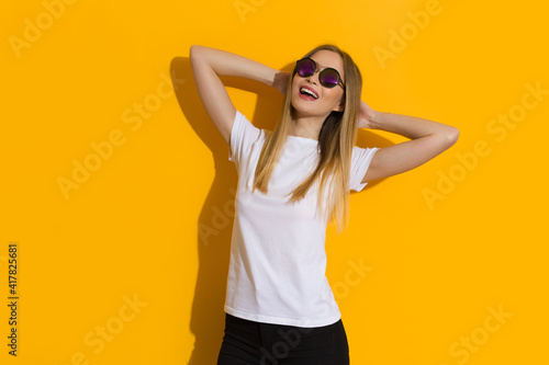 Relaxed Young Blond Woman In White Shirt And Sunglasses Is Posing In The Sunlight Against Yellow Wall