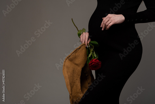 Close-up of a pregnant belly. Black bodycon dress. Hat and rose in hands. Grey background.