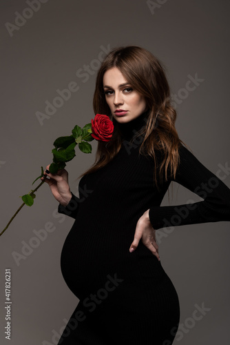 Pregnant girl in black tight dress. With a rose in my hand. Hand on the waist.  Grey background.