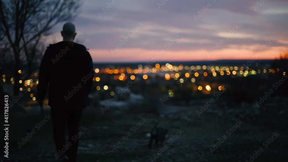 Un hombre joven pasea a su perro en la cima de una montaña, de fondo se ven las luces de la ciudad de noche y la puesta del sol