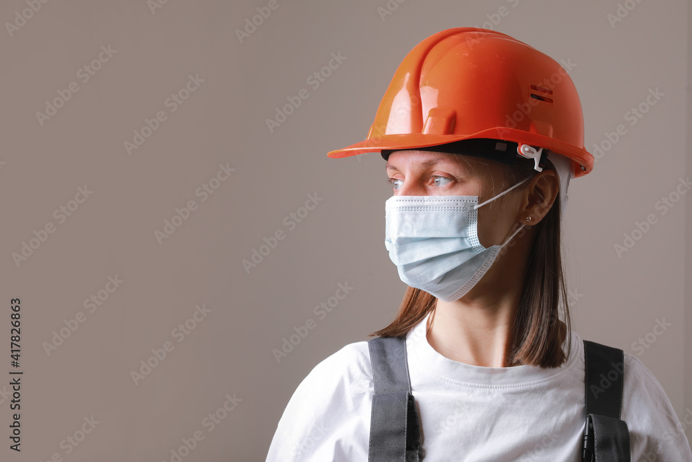 Pretty woman worker in medical mask and orange construction helmet on a gray background. Occupational safety and health specialist with face mask for coronavirus covid-19 Pretty woman worker in medical mask and orange construction helmet on a gray background. Occupational safety and health specialist with face mask for coronavirus covid-19