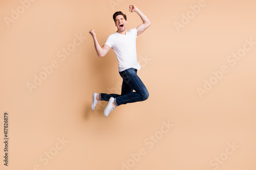 Full size profile photo of hooray brunet man jump yell wear t-shirt jeans sneakers isolated on beige color background