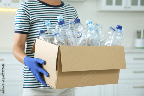 Woman holding cardboard box with used plastic bottles indoors, closeup. Recycling problem © New Africa