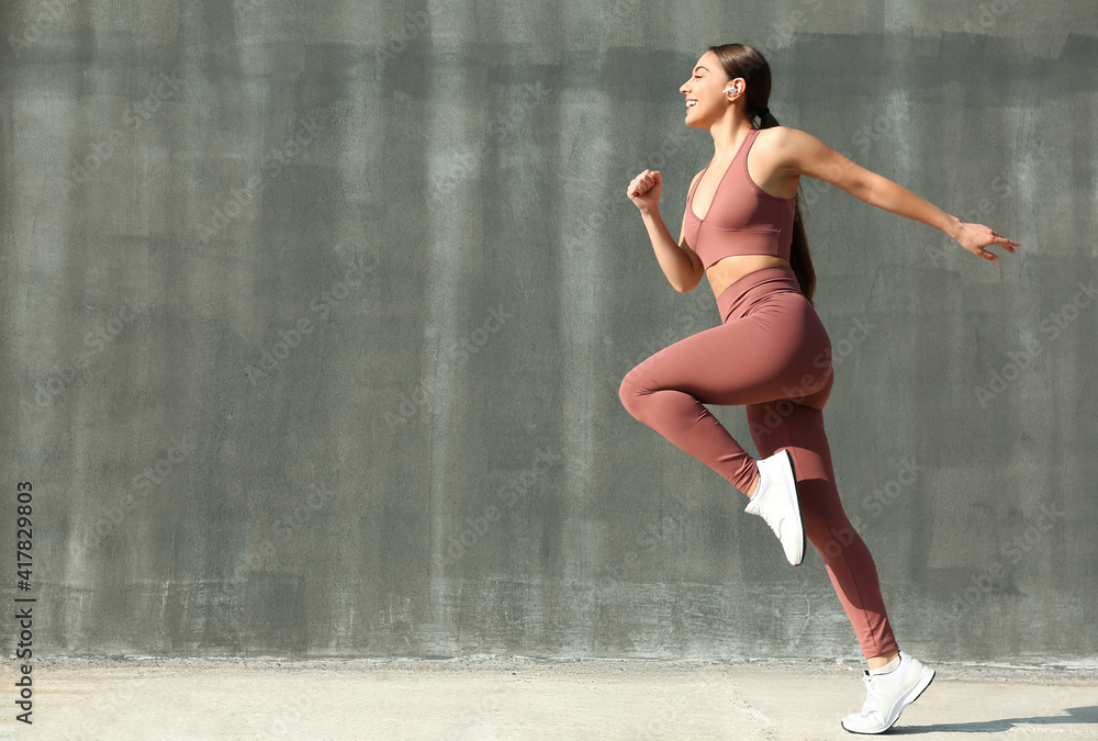 Happy young woman in stylish sports wear jumping near grey wall. Space for text