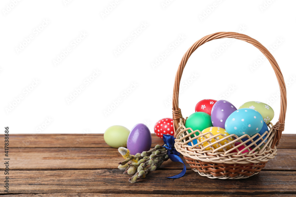 Colorful Easter eggs in wicker basket and willow branches on wooden table against white background. Space for text