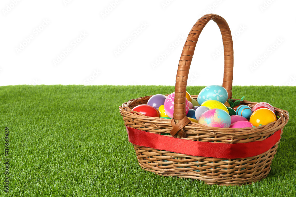 Wicker basket with Easter eggs on green grass against white background. Space for text