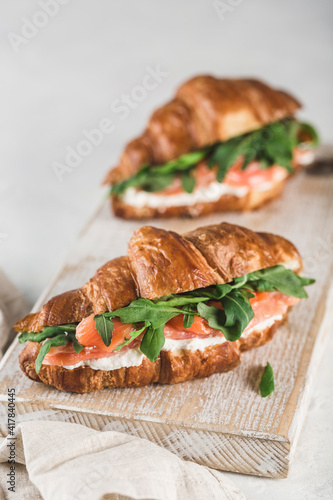 Two croissant sandwich with salmon, ricotta cheese and arugula on a wooden board with a napkin on a light background.