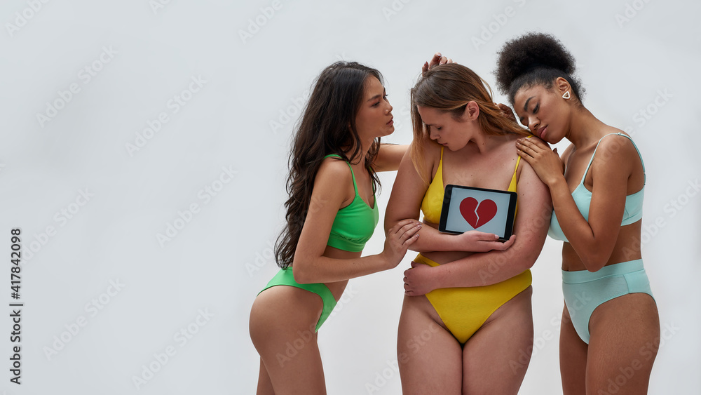 Two young women supporting other curvy woman wearing colorful underwear looking shy, holding tablet pc with red broken heart shape on screen, posing together over light background