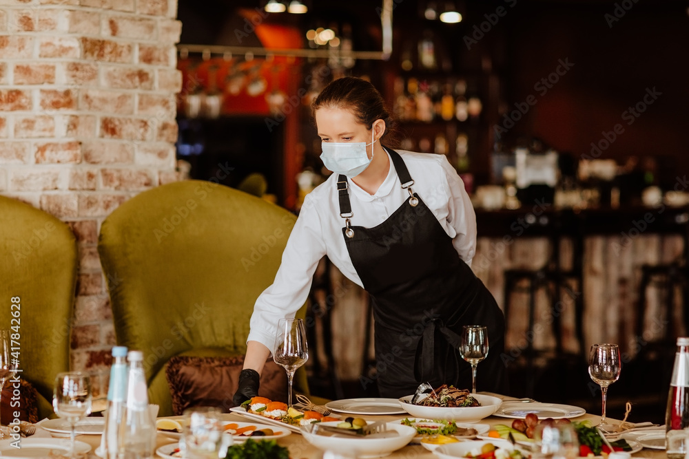 Young female waiter in uniform with medical mask and gloves serving in the restaurant. Copy space. Young female waiter in uniform with medical mask and gloves serving in the restaurant. Copy space.