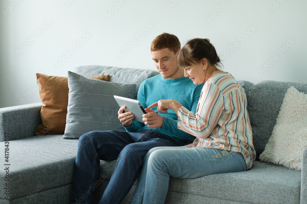 son teaching his mother to use tablet. older people using technology. Cheerful elderly woman sitting on the sofa next to his adult son son teaching his mother to use tablet. older people using technology. Cheerful elderly woman sitting on the sofa next to his adult son