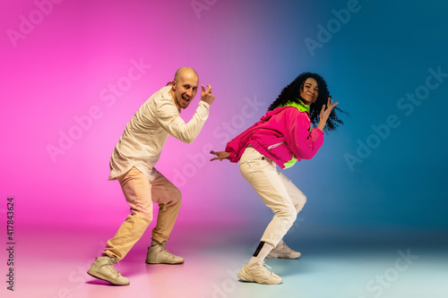 Team. Stylish sportive caucasian couple dancing hip-hop on colorful gradient background at dance hall in neon light. Youth culture, movement, style and fashion, action. Fashionable bright portrait.