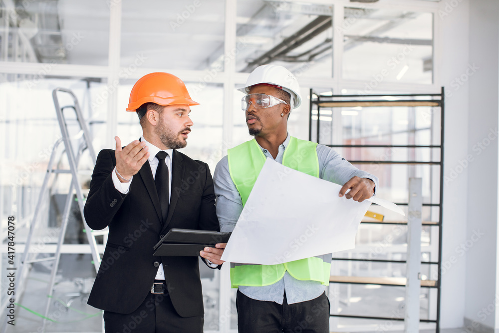 African and caucasian men in protective helmets using digital tablet and blueprints while controlling working process on construction. Cooperation concept. African and caucasian men in protective helmets using digital tablet and blueprints while controlling working process on construction. Cooperation concept.