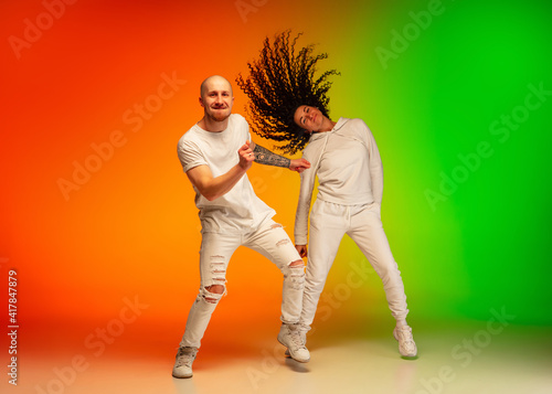 Stylish sportive caucasian couple dancing hip-hop on colorful gradient background at dance hall in neon light. Youth culture, movement, style and fashion, action. Fashionable bright portrait.