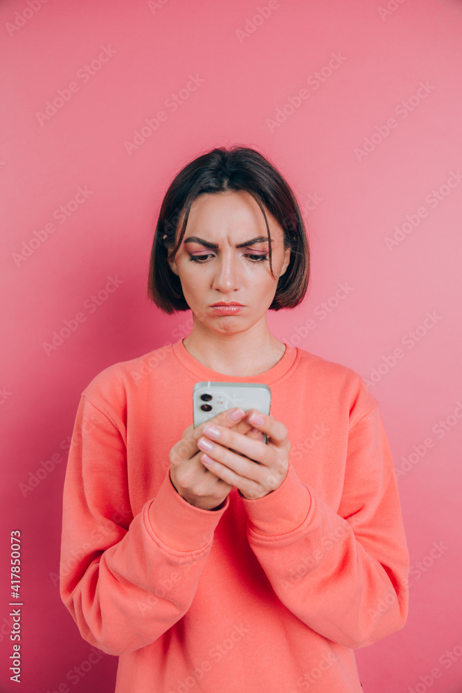 Young woman is feeling sad unhappy angry while reading sms using her mobile phone, over pink background Young woman is feeling sad unhappy angry while reading sms using her mobile phone, over pink background