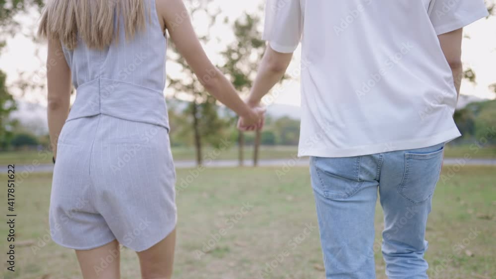 View from behind of cheerful couple holding hands while talking a walk together through green natural park, happiness people lifestyle, young age relationship goal, romantic couple free from Covid-19 View from behind of cheerful couple holding hands while talking a walk together through green natural park, happiness people lifestyle, young age relationship goal, romantic couple free from Covid-19