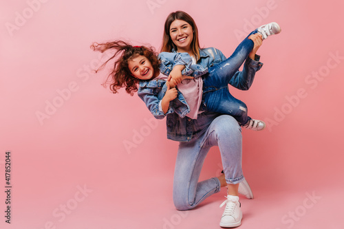 Smiling young woman playing with daughter. Studio shot of happy mom and preteen kid in denim attire.