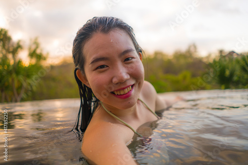 young woman relaxed at luxury resort infinity pool - happy and cheerful Asian Korean girl taking selfie photo in bikini enjoying jungle view at hotel swimming pool during summer holidays