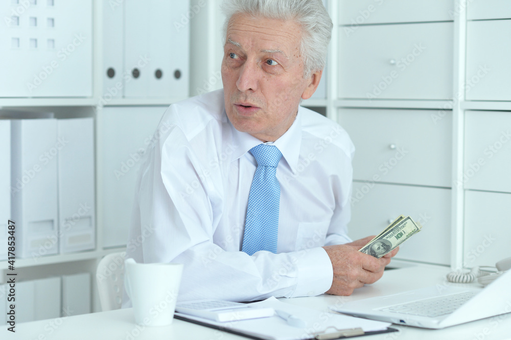 Senior doctor sitting at table with laptop and holding money Senior doctor sitting at table with laptop and holding money