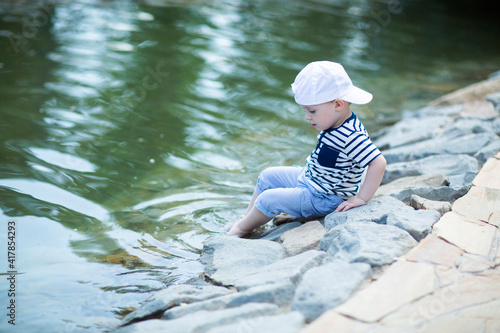 Fototapeta Naklejka Na Ścianę i Meble -  Baby in white cap sitting by the pond touches cold water with his leg and admires