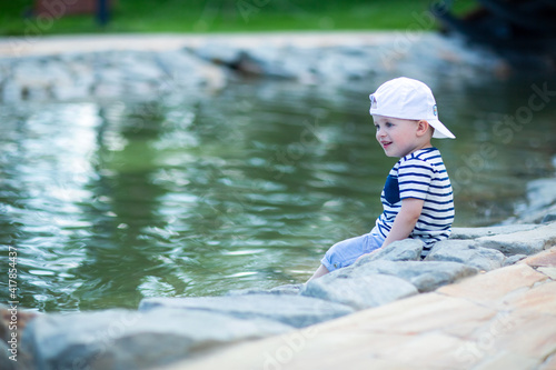Fototapeta Naklejka Na Ścianę i Meble -  little naughty boy is happy and smiling, sitting on the edge of a pond in a city park, wearing a cap turned back