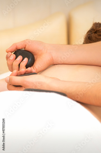 Hands of masseur hold black hot stones massaging back of a young adult woman in spa salon