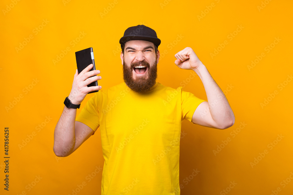 Photo of amazed bearded man celebrating victory and holding tablet over yellow background Photo of amazed bearded man celebrating victory and holding tablet over yellow background
