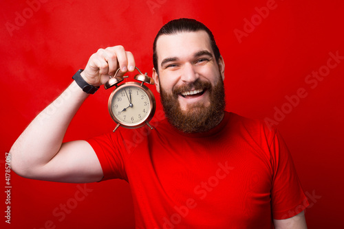 Photo of cheerful young bearded man holding alarm clock over red background