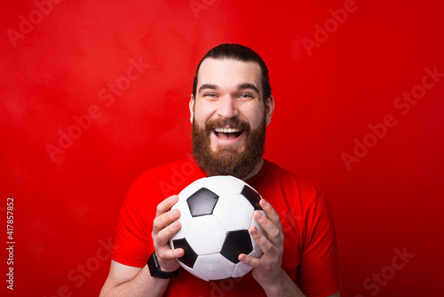 Smiling young bearded man standing over red background and holding soccer ball © Vulp