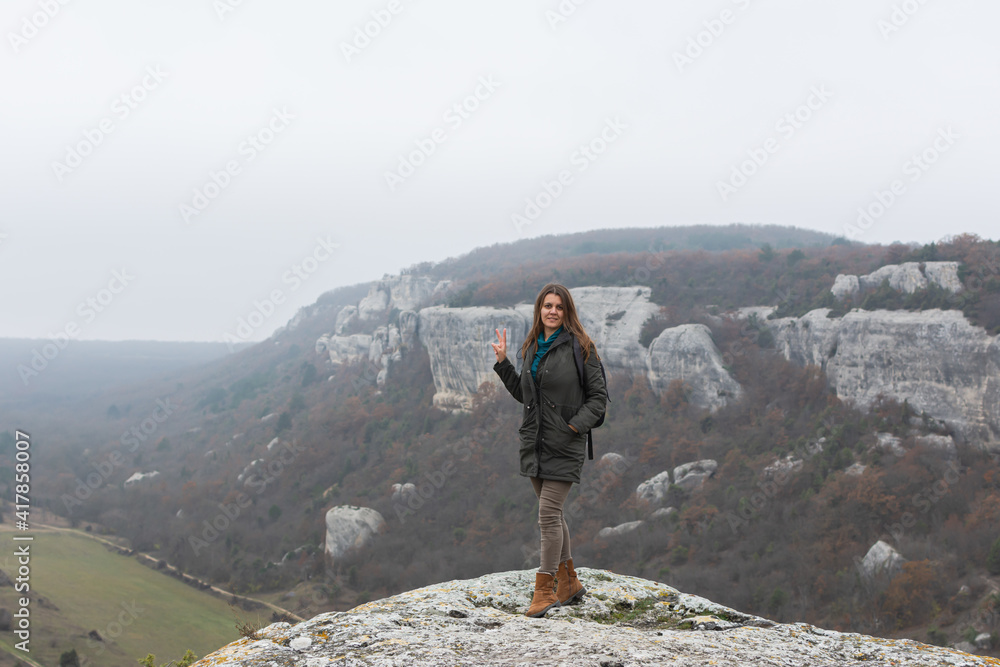 Obraz premium A young dark-haired woman on the background of an autumn landscape. Cloudy gray foggy view from the mountain. A happy smiling girl in a green coat. Natural background, concept of hiking, ecotourism
