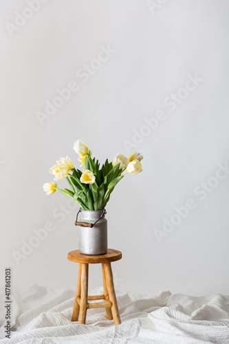 gelb weiße Tulpen als Blumenstrauß in einer vintage Zink Milchkanne arrangiert auf einem braunen Holzhocker und lockerem Stoff vor grauem Hintergrund