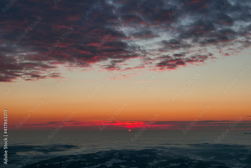 A sunrise seen from snow-capped peak of Schoeckl in Austrian Alps. The sky is bursting with orange and pink. Distant view on Graz, shrouded with fog. Winter wonderland. Day break. Calmness