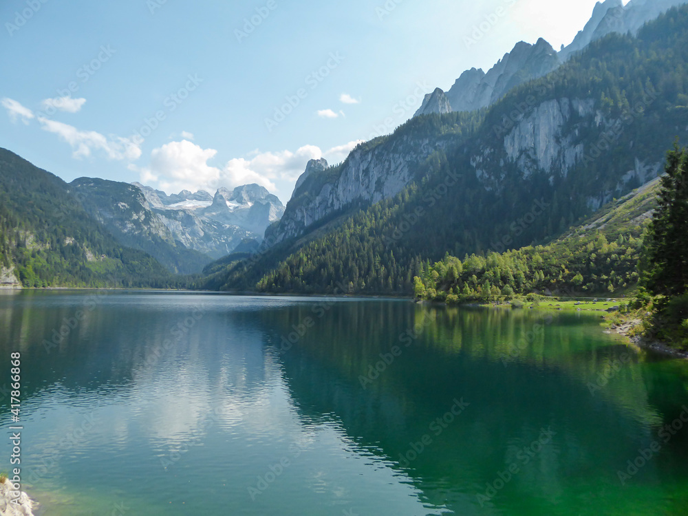 Panoramic view on Gosau lake, with Dachstein glacier in the back in Austrian Alps. The lake is surrounded by high mountains, overgrown with tall trees. Sun reflects on the surface. Serenity  and calm
