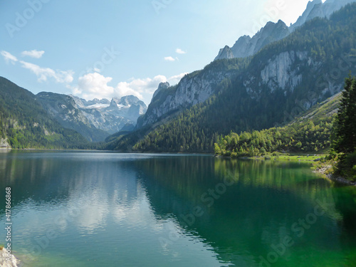 Panoramic view on Gosau lake, with Dachstein glacier in the back in Austrian Alps. The lake is surrounded by high mountains, overgrown with tall trees. Sun reflects on the surface. Serenity and calm