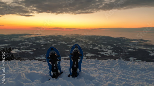 A pair of snow shoes stuck into powder snow at the peak of Schoeckl in Austrian Alps, with the view on sun rising above the horizon. The sky is bursting orange. Winter wonderland. Day break. City view