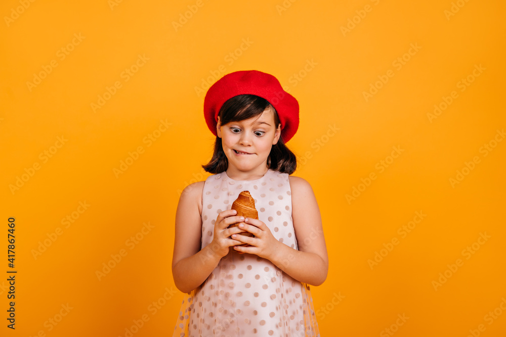 Studio shot of little girl holding croissant. Brunette kid in french beret expressing amazement on yellow background.