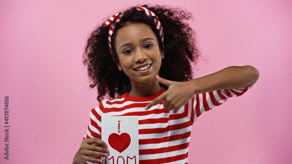 happy african american girl pointing with finger at greeting card with i love mom lettering isolated on pink happy african american girl pointing with finger at greeting card with i love mom lettering isolated on pink