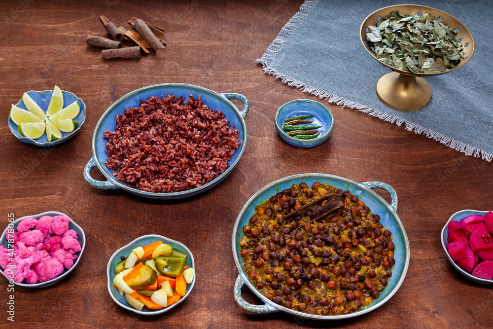 Set of plates and bowls on wooden table. Black chickpea (Kala chana) curry and red rice, vegetable pickles. Vegan meal. Set of plates and bowls on wooden table. Black chickpea (Kala chana) curry and red rice, vegetable pickles. Vegan meal.